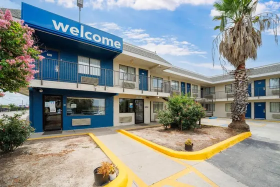 White motel with a blue Welcome sign, palm, and pink flowers
