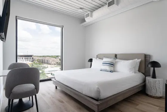 White bedroom with a wood bed, gray chair, and city window